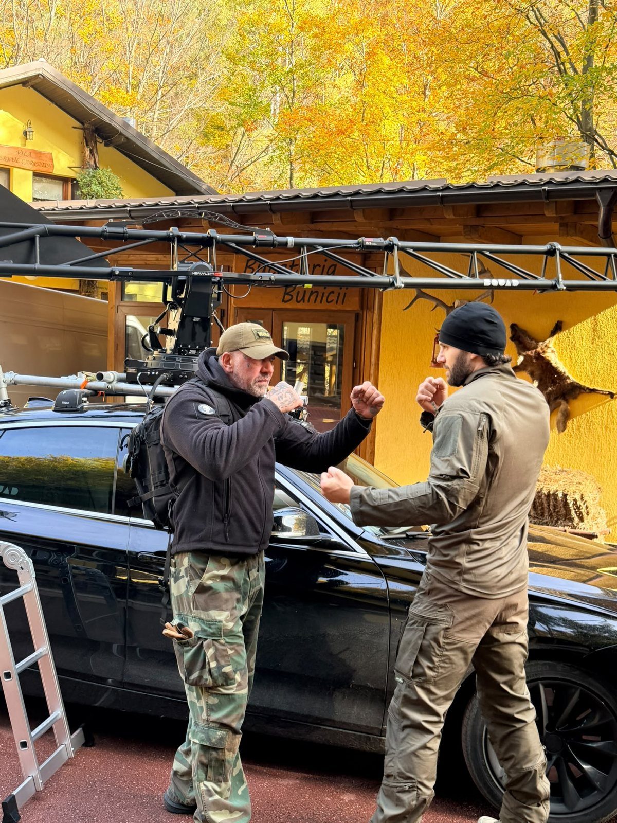 Two people rehearsing fight choreography beside a car and camera rig on a film set